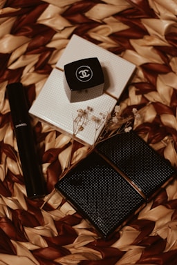An arrangement of Chanel cosmetic products on a woven textured background. There is a black cylindrical tube, likely a lipstick or mascara, alongside a small black square container with the Chanel logo. A couple of white, textured, square items and some delicate dried flowers are also present.