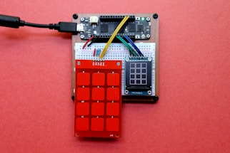 A small electronic circuit setup featuring a breadboard connected to a microcontroller and sensors. There is a red keypad sensor with numbered squares and a small LCD display adjacent to it, all placed on a pink background. Various colored jumper wires link the components together.