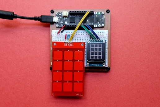 A small electronic circuit setup featuring a breadboard connected to a microcontroller and sensors. There is a red keypad sensor with numbered squares and a small LCD display adjacent to it, all placed on a pink background. Various colored jumper wires link the components together.