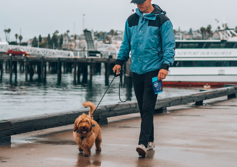 Hombre con chaqueta azul y pantalones negros de pie junto al perro marrón de pelo largo en el muelle durante