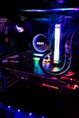 A close-up view of a custom-built computer's internal components, featuring a prominent liquid cooling system from NZXT. Colorful RGB lighting illuminates the interior, giving it a futuristic look. The setup includes a graphics card and various LED-lit cables, all enclosed within a transparent case.