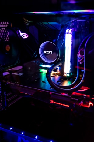 A close-up view of a custom-built computer's internal components, featuring a prominent liquid cooling system from NZXT. Colorful RGB lighting illuminates the interior, giving it a futuristic look. The setup includes a graphics card and various LED-lit cables, all enclosed within a transparent case.