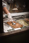 Artisan gelato being carefully scooped from a display case with beach waves reflected in the glass.