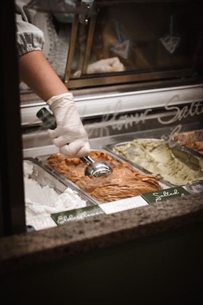 A gloved hand uses a metal scoop to serve gelato from a display case containing several flavors. The foreground shows tubs of white, brown, and green gelato, each labeled with handwritten signs.