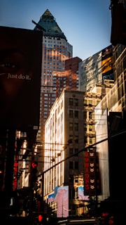 Tall buildings with billboards and advertisements line a bustling city street. The light reflects off the glass windows of the skyscrapers. Signs for Broadway musicals and other promotions are prominently displayed.