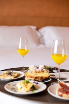 A breakfast tray is placed on a bed, featuring two glasses of orange juice and an assortment of breakfast foods such as croissants, toast, omelets, and pastries. The bed has a white duvet and pillows, creating a cozy and inviting atmosphere.