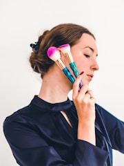 A person with a side profile is holding two bright makeup brushes with multicolored handles and fluffy pink bristles against a plain background. The individual has long hair tied back and is wearing a dark satin-like blouse.