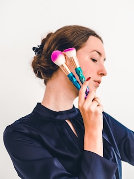 A person with a side profile is holding two bright makeup brushes with multicolored handles and fluffy pink bristles against a plain background. The individual has long hair tied back and is wearing a dark satin-like blouse.