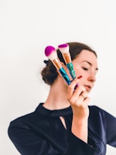 A person holding two makeup brushes with vibrant pink bristles and intricately designed handles. The background is plain, highlighting the focus on the brushes. The person's face is partially out of focus, drawing attention to the objects in hand.