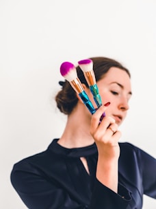 A person holding two makeup brushes with vibrant pink bristles and intricately designed handles. The background is plain, highlighting the focus on the brushes. The person's face is partially out of focus, drawing attention to the objects in hand.
