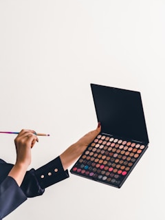A person is holding a makeup palette filled with a variety of colorful eyeshadows in one hand and a makeup brush in the other. The palette contains multiple shades ranging from neutrals to vibrant colors like pink and blue. The background is a plain light color, and the person is wearing a dark sleeve with metallic buttons.