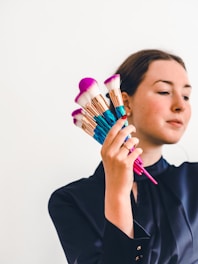 A person holding a set of colorful makeup brushes against a plain background. The brushes have golden ferrules and vibrant pink and blue handles, while the bristles are white with pink tips. The person is wearing a dark blue top.