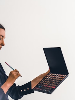 A person holds an open makeup palette with a wide variety of colors and is using a thin brush to apply eyeshadow or makeup. The person is wearing a dark long-sleeved top, and the background is minimal and light-colored, providing contrast.