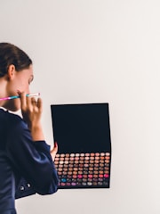 A person holding a large makeup palette filled with various colorful eyeshadows or makeup powders. The individual appears to be using a makeup brush while looking at or selecting a color.