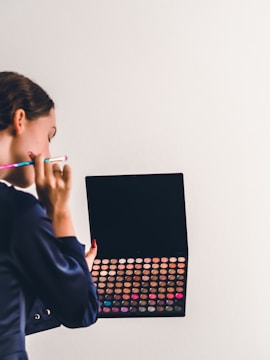 A person holding a large makeup palette filled with various colorful eyeshadows or makeup powders. The individual appears to be using a makeup brush while looking at or selecting a color.