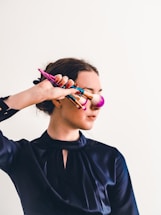 A person holds several colorful makeup brushes in front of their face, wearing a dark satin-like blouse against a plain light background. The pink brush head lightly touches their cheek.