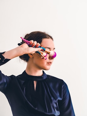 A person holds several colorful makeup brushes in front of their face, wearing a dark satin-like blouse against a plain light background. The pink brush head lightly touches their cheek.