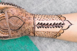 A forearm displays intricate henna designs featuring a combination of floral patterns, geometric shapes, and bold vine motifs. The design is detailed and symmetrical, covering the back of the hand and extending along the forearm.