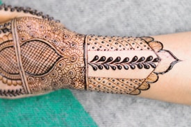 A forearm displays intricate henna designs featuring a combination of floral patterns, geometric shapes, and bold vine motifs. The design is detailed and symmetrical, covering the back of the hand and extending along the forearm.