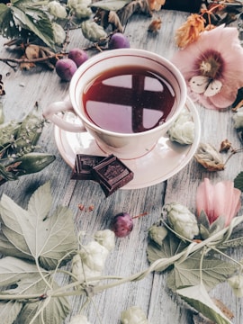 A porcelain cup filled with tea sits on a saucer, surrounded by small dark chocolate pieces. Around the cup are various natural elements, including leaves, a butterfly, flowers, and some small fruits, all arranged on a rustic wooden surface.