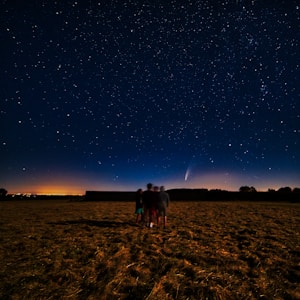 A group of people standing in an open field under a night sky filled with stars. A bright comet with a visible tail is prominently featured among the stars, creating a striking celestial scene. The ground is grassy and dimly lit, while the horizon glows softly with light from a distant city or sunset.