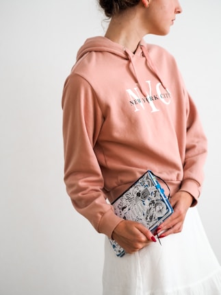 A person wearing a peach-colored hoodie with 'New York City' written on it is holding a patterned notebook. The person has their hair tied back and is looking to the side. The background is plain and light-colored.
