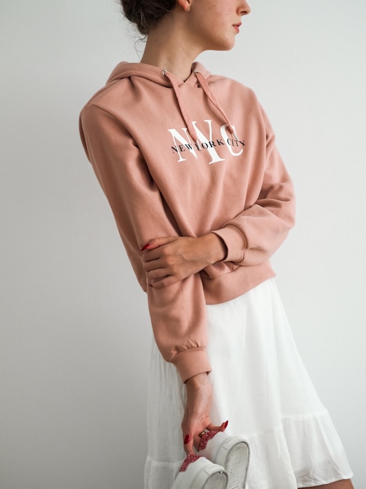 Person wearing a light brown hoodie with 'New York City' printed on it and a white skirt, holding a pair of white sneakers.