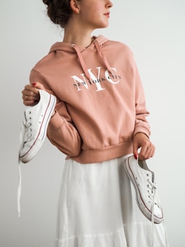 A person wearing a peach-colored hoodie with 'New York City' printed on it holds a pair of white sneakers, one in each hand. The person is also wearing a white skirt and looking to the side with a neutral expression.