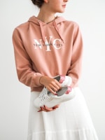 A person wearing a peach-colored hoodie with 'New York City' written on it is holding a pair of white sneakers with red glittery accents. The person is also dressed in a white skirt, and their nails are painted red.