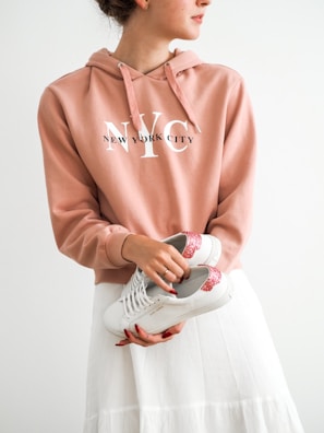 A person wearing a peach-colored hoodie with 'New York City' written on it is holding a pair of white sneakers with red glittery accents. The person is also dressed in a white skirt, and their nails are painted red.