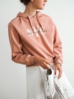 A person is wearing a peach-colored hoodie with 'New York City' printed on it, paired with a white skirt. They are holding a pair of white sneakers with red accents. The person has their hair tied back and is wearing a small earring.