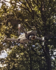 A drone with a sleek, metallic design flies amidst a natural setting. The rotors are visible, and the background features lush green trees and soft sunlight filtering through the leaves.