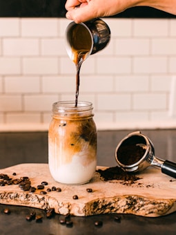 A hand pours espresso from a small metal pitcher into a glass jar filled with milk and ice. The jar sits on a rustic wooden board surrounded by scattered coffee beans. An espresso portafilter filled with coffee grounds lies nearby, adding a touch of authenticity and warmth to the scene. The background consists of white subway tiles, contributing to a clean and minimalist aesthetic.