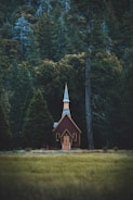A small, rustic church with a pointed steeple situated in a lush forest clearing. The church is wooden with dark red walls and light trim, blending harmoniously with the surrounding tall trees and dense forest vegetation.