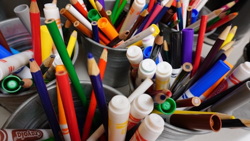 A variety of colored pencils and markers are arranged in metal containers. The collection includes a range of vibrant colors such as red, green, blue, yellow, and more. Some pencils are sharpened, while others are dull. The markers have both thick and thin tips, and there are visible brand markings like Crayola.