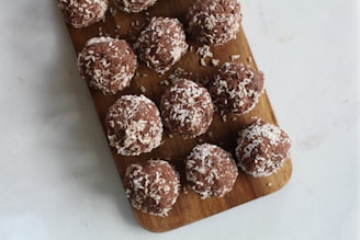 A wooden cutting board holds multiple coconut-covered chocolate balls arranged neatly. The tabletop appears to be light-colored, possibly marble or quartz, enhancing the contrast with the dark brown board and chocolate treats.