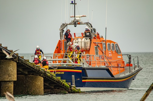 A large orange lifeboat is stationed near a concrete structure in the water. Several crew members wearing red and black uniforms and helmets are aboard the vessel. The lifeboat belongs to a search and rescue organization and appears to be preparing for or returning from a mission. The background features a calm sea under a cloudy sky. A gull is flying near the structure on the left side of the image.