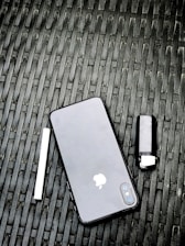 A smartphone with a black back cover featuring an apple logo is placed on a woven black surface. Next to it, on the left, is a white cylindrical object and on the right is a dark-colored lighter.