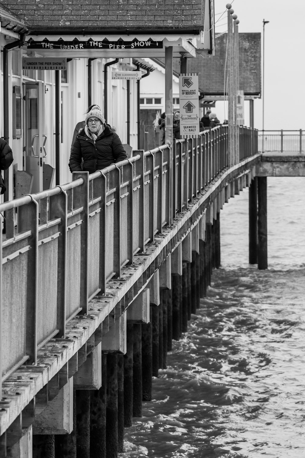 grayscale photo of woman in black jacket standing on bridge
