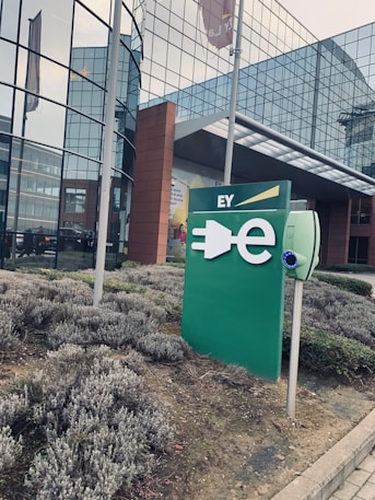 A modern office building with large glass windows. In front of the building, there is a green sign with a plug symbol and the letters 'EY'. Surrounding the area are neatly trimmed bushes.