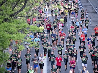 A large group of runners participates in a marathon on a city street lined with green trees. The participants wear various colorful athletic clothing, with many sporting numbered race bibs. The atmosphere is lively, with runners of diverse ages moving at different paces.