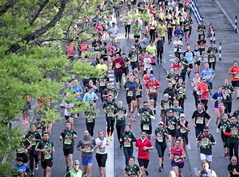 A large group of runners participates in a marathon on a city street lined with green trees. The participants wear various colorful athletic clothing, with many sporting numbered race bibs. The atmosphere is lively, with runners of diverse ages moving at different paces.