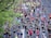 A large group of runners participates in a marathon on a city street lined with green trees. The participants wear various colorful athletic clothing, with many sporting numbered race bibs. The atmosphere is lively, with runners of diverse ages moving at different paces.