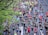 A large group of runners participates in a marathon on a city street lined with green trees. The participants wear various colorful athletic clothing, with many sporting numbered race bibs. The atmosphere is lively, with runners of diverse ages moving at different paces.