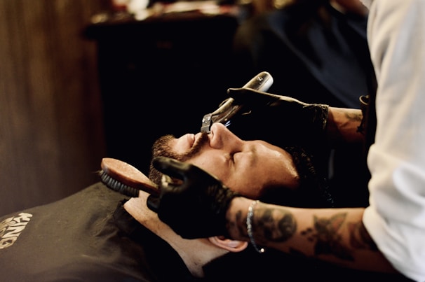 A person is lying back in a barber's chair while another person, likely a barber, is shaping their beard with a straight razor. The person in the chair has eyes closed, appearing relaxed. The barber wears gloves and has tattooed arms. A brush is also being used to groom the beard.
