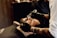 A person is lying back in a barber's chair while another person, likely a barber, is shaping their beard with a straight razor. The person in the chair has eyes closed, appearing relaxed. The barber wears gloves and has tattooed arms. A brush is also being used to groom the beard.