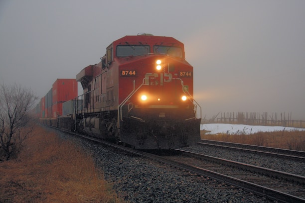 A freight train with bright headlights travels through a foggy landscape, carrying numerous shipping containers. The surroundings include dry grass and tracks leading into the distance, with an overcast and moody sky.