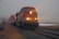 A freight train with bright headlights travels through a foggy landscape, carrying numerous shipping containers. The surroundings include dry grass and tracks leading into the distance, with an overcast and moody sky.