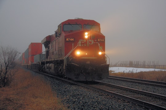 A freight train with bright headlights travels through a foggy landscape, carrying numerous shipping containers. The surroundings include dry grass and tracks leading into the distance, with an overcast and moody sky.