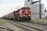 A freight train with a red locomotive is traveling on railway tracks beside large, industrial silos and structures. The sky is overcast, and the area appears to be part of an industrial zone.